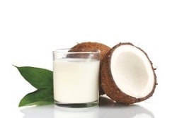 Auf dem Bild ist rechts von einem Glas mit kokosnussmilch geüllt, eine halb aufgeschnittene Kokosnuss zu sehen. Dahinter befindet sich noch eine ganze Kokosnuss. Links vom Glas ist das Bild mit einem grünen Blatt verziert. Der Hintergrund ist weiß.