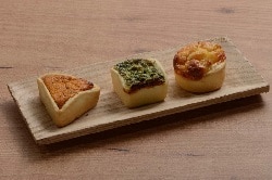 Auf dem Foto sind die drei verschiedenen Mini-Quiche-Sorten des Herstellers Maitre Pierre zu erkennen. Sie sind auf einem länglich quadratischem Holzteller auf einem Holzfußboden. Links befindet sich ein dreieckiges Stück, in der Mitte ein quadratisches und rechts eine runde Mini-Quiche.