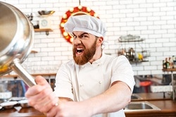 Ein in weiß gekleideter Koch ist frustriert über den Personalmangel in der Gastronomie und hebt eine silberne Pfanne mit beiden Händen hoch um zuzuschlagen. Er hat den Mund weit geöffnet. Im Hintergrund ist eine Küche zu sehen.