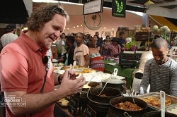 Auf dem Bild ist der Food Experte Andrew Fordyce zu sehen, der auf einem Markt in Südafrika neue Gerichte und Snack testet. Er trägt ein rotes Shirt, einen Sonnenbrille auf dem Kopf und hat eine Tragetasche quer über der Brust. Vor ihm auf dem Tisch sind Kochtöpfe mit Fleisch, aber auch Beilagen wie Reis aufgestellt. Hinter dem Tisch steht ein Verkäufer in einem grau gestreiftem Langarm-Shirt.
