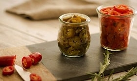 Zwei kleine Gläser nebeneinander stehend , gefüllt mit geschnittenen Chilis. Ein Glas enthält grüne, das andere rote Chilis. Links daneben ist eine angeschnittene rote Chili zu sehen. Messer und Abschnitt liegen neben der geschnittenen Chili. 