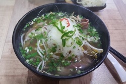Auf dem Bild erkennt man das vietnamesische Gericht Pho, welches aus Reisnudeln besteht. Dazu werden Lauch und verschiedene Gemüse in einer Brühe serviert. Das Essen befindet sich in einer schwarzen Schale auf einem Holztisch.