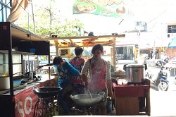 Auf dem Foto erkennt man einen Stand in Asien, an dem eine Frau und zwei Männer kochen. Die Frau, die zur Kamera gewandt ist,. trägt über dem weißen Langarm-Shirt eine rosa Schürze und kocht im Wok. Neben ihr ist ein Junge in einem blauen Shirt. Vor ihm steht ebenfalls ein Wok, der aber nicht benutzt wird. Dahinter ist der Stand zur Straße, an dem ein anderer Junge steht um für die Leute zu holen. Rechts auf dem Bild erkennt man einen Tisch auf dem ein Reiskocher steht.