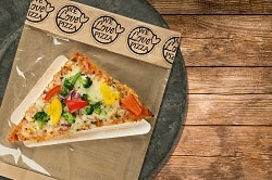Auf dem Foto ist eine dreieckige Pizza abgebildet. Es ist der Point Snack vom Pizzahersteller Point of Food. Die Pizza ist belegt mit Gemüse aller Art wie gelber Paprika, roter Paprika, Zwiebeln und Brokkoli. Diese Variante des Point Snacks nennt sich Verdura. Die Pizza ist auf einem dreieckigen Pappslice in einer Verpackung zu sehen. Man erkennt, dass diese Pizza sich in einer Verpackung befindet. Oberhalb der braunen Tütenverpackung steht in schwarzer Schrift "We love Pizza" mehrmals in einem runden KReis geschrieben. Die verpackte Pizza befindet sich auf einem dunkel-grauen Teller auf einem Holztisch.