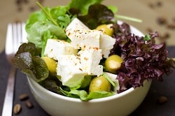 Auf dem Foto ist ein gemischter Salat zu erkennen. Neben den Salatblättern erkennt man grüne Oliven und Feta-Käse in der weißen Schüssel. Links von der Schale befindet sich eine Gabel.