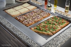 Auf dem Bild erkennt man drei verschiedene Pizzen, die auch jeweils anders geschnitten sind. Vorne rechts ist eine Pizza in P-Form geschnitten, welche mit Rucola und Tomaten belegt ist. In der Mitte ist eine Pizza zu sehen, die in sechs gleich große Quadrate geteilt ist. Die Pizza ist belegt mit Thunfisch und roten Zwiebeln. Die letzte und hinterste Pizza hat das selbe Schnittmuster wie die zweite pizza, ist jedoch mit ausgiebig viel Käse belegt. Die Pizzen befinden sich in einer Kantine oder in einem Bistro.