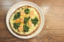 Auf dem Foto ist eine Pizza aus dem High Protein Teig des Herstellers Point of Food zu sehen, die mit einer gelben Sauce, Lachs und Spinat belegt ist. Die Pizza ist auf einem weißen Teller auf einem Holztisch. 