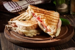 Ein gegrilltes Sandwich belegt mit Tomate, Schinken und Käse ist auf einem Holzteller auf einem Holztisch. Das Sandwich ist diagonal geschnitten, sodass zwei Dreiecke auf dem Teller sind. Anhand der braunen Streifen und des knusprigen Toastes erkennt man, dass das Brot gegrillt wurde. Der Käse zwischen den Scheiben ist geschmolzen und verläuft.