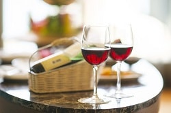 Hotellkongress_Auf dem Bild sind zwei mit Rotwein gefüllte Weingläser zu sehen, die auf einem Mamortisch stehen. im Hintergrund liegt die Weinflasche des Weines im Glas in einen hellbraunen Weidenkorb.