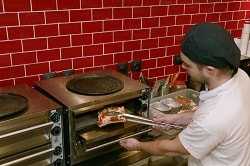 Auf dem Foto ist ein Backofen zu sehen, der von einem Koch bedient wird. Mit Hilfe einer Zangee legt er ein Stück Pizza in den Ofen, um dieses aufzubacken. Der Ofen ist quadratischen und aus Stahl. Der Koch trägt ein weißes Shirt und ein schwarzes Tuch auf dem Kopf. Mit der linken Hand öffnet er den Ofen, mit der rechten bedient er die Zange für die Pizza. Die Kacheln hinter dem Ofen sind alle rot.