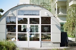 Man erkennt ein Burger Restaurant, welches Bio Burger serviert. Über der Tür steht Burger und links von der Tür das englische Wort für Bio, welches organic heißt. Der Laden sieht aus wie ein Container, man sieht ihn von vorne. Er ist halbrund und grau mit großen Fenstern an der Vorderseite, das heißt da wo sich die Tür befindet. 