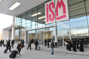 Der Eingang der Kölnmesse, wo die ISM stattfindet, ist auf dem Foto abgebildet. Über der Eingangstür hängt auf weißen Hintergrund in pinker Schrift der Schriftzug ISM. Die ISM ist die internationale Fachmesse für Süßwaren.