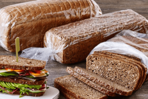 Auf dem Bild erkennt man das abgepackte Brot in einem Brotschlauch von Kohberg. Links von den drei Brote, wovon zwei abgepackt und eins geöffnet ist, befindet sich ein Belegbeispiels mit Salat, Gemüse und weiterem Belag.