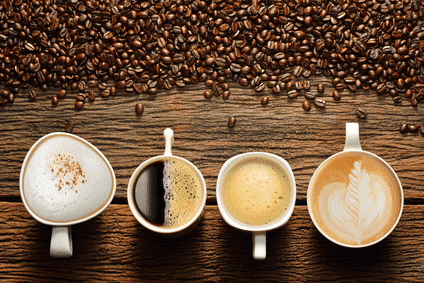 Vier verschiedene Variationen von Kaffee sind aus der Vogelperspektive zu sehen. Von links ist es ein Cappuccino, ein Americano, also Kaffee schwarz, ein Kaffee Crema und ein Milchkaffee. Die Kaffees stehen auf Holzboden und oberhalb des Bildes sind ganze Kaffeebohnen verstreut.