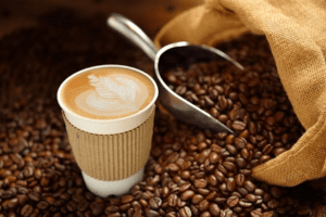 Ein Kaffee in einen Pappbecher steht zwischen vielen Kaffeebohnen, die gerade aus einem Sack Kafee raus fallen. Außerdem befindet sich neben dem Kaffeebecher eine silberne Schaufel für die Bohnen.