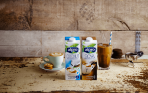 Die Milchalternativen Soya-Milch und Kokosnuss-Milch stehen vor einer Steinwand. Neben der Kokosnuss-Milch befindet sich ein Kakaoin eine Latte Macchiato Glas mit einem Strohalm, links von der Soya-Milch ist ein Cafe Crema zu sehen.