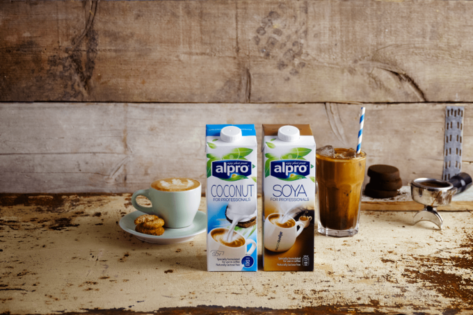Die Milchalternativen Soya-Milch und Kokosnuss-Milch stehen vor einer Steinwand. Neben der Kokosnuss-Milch befindet sich ein Kakaoin eine Latte Macchiato Glas mit einem Strohalm, links von der Soya-Milch ist ein Cafe Crema zu sehen.