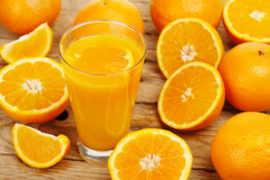 Ein Glas ausgepresster Orangensaft steht in der Mitte von vielen Orangenhälften, aber auch ganzen Orangen auf einem Holztisch.