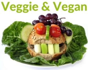 Dunkles Brötchen auf einem Salatblatt, als Gesicht gestaltet. Gurken Ohren, Tomaten Augen, Weintrauben Haare, und Gemüse Zähne. Das Bild hat die grüne Überschrift, Veggie und Vegan