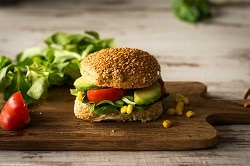 Das Bild zeigt einen Burger auf einer Holzplatte auf einem Holztisch. Der Burger besteht aus einem dunklen Brötchen, Salat, Tomate und Avocadohälften. Im Hintergrund erkennt man Salat, der zur Dekoration dient.