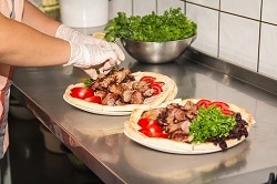 Zwei Hände die weiße Handschuhe an haben und einen dicken Pizza Teig belegen. mit gebratenen großen Fleischstücken und grünem Salatblatt.