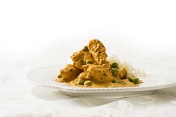 Auf dem hellen Bild ist eine indische warme Mahlzeit auf einem weißen Teller zu sehen. Rechts auf dem Teller befindet sich ein wenig Reis. Die gelbe-Soße mit grünen Erbsen und vielen und großen Stücken zartem Hühnchenfleisch steht im Fokus des Bildes. Der Hintergrund ist weiß.