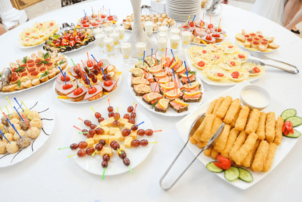 Auf dem Tisch sind mehrere Teller mit Snacks und kleinen Häppchen gedeckt. Links befinden sich auf dem weißen Teller Spieße mit Käse und Weintrauben. Rechts davon erkennt man Fischstäbchen, die mit einer Zange, die an der linken Ecke des eckigen Tellers ist, vom Gast genommen werden sollen. Außerdem erkennt man dahinter mehrere Variationen von Butterbroten, mit Wurst oder Käse belegt und mit Obst verziert. In den Broten, ebenso wie in den Schokoladen- und Vanillebällchen links, stecken kleine Spieße zum aufgreifen der Snacks. In der MItte des Tisches ist ein weißes Standbein zu sehen, möglicherweise das eines Kerzenständers. Um dieses Bein herum sind Kurzegläser gefüllt mit einer Art Pudding oder Creme, die den Nachtisch und die süße Versuchung darstellen soll.