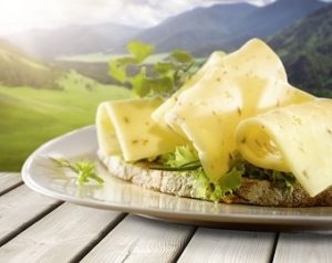 Auf dem Foto ist ein BUtterbrot mit drei eingerollten Käsescheiben zu sehen. Der Käse ist der neue würzige Käse von Frischpack. Auf dem Käsebrot befindet sich unter dem Käse ebenfalls noch Salat. Das belegte Brot liegt auf einem weißen Teller, auf einem hell-grauen Holztisch. Im Hintergrund ist eine Landschaft aus Wiesen und Bergen zu erkennen. Die Wiesen sind grün und die Berge verschimmen in einer weiß-grau und bläulichen Farbe.