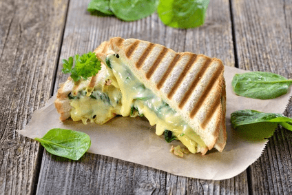 Auf einem Pergamentpapier ist ein heißes, gegrilltes Sandwich zu erkennen. Der Snack ist mit Spinat und Käse belegt. Durch das Grillen ist der Käse geschmolzen und das Sandwich hat Grillstreifen bekommen.