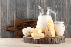 Eine Glaskanne mit Milch, Frischkäse, Quark im Glas und zwei verschiedene Sorten Käse liegen auf einer Holzplatte. Die Milchprodukte stehen vor einer Holzwand und einem dunklen Schneidebrett auf einem hellen Holzboden.