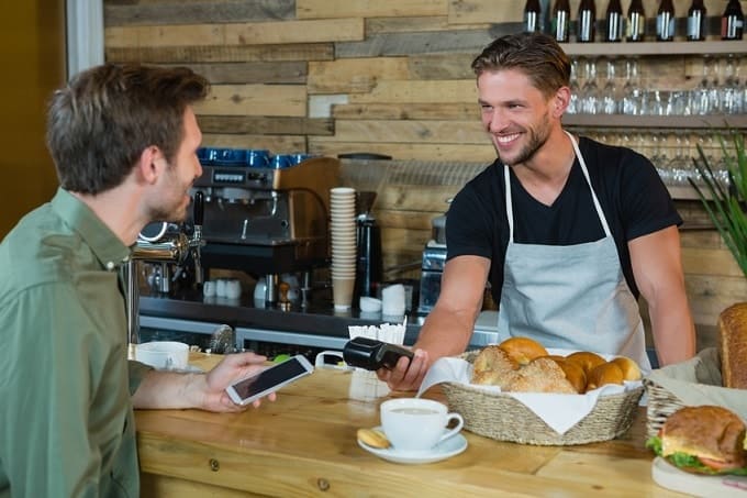 Ein Mann mit einem Smartphone in der Hand und ein Mann mit einem NFC Zahlungsgerät in der Hand. Sie stehen in einem Café mit Backwaren auf dem Tresen.