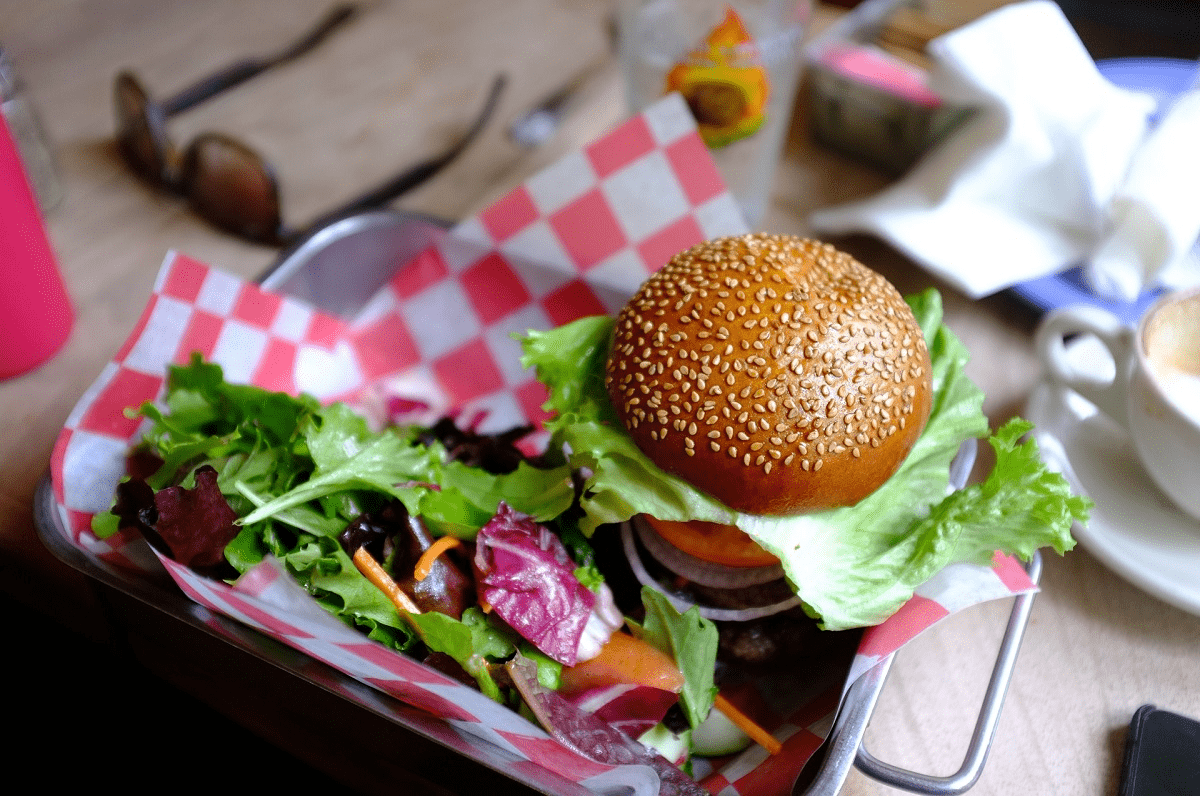 Auf dem Foto ist ein Burger mit einer Salatbeilage in einem Metallschüssel zu erkennen. Unter dem Burger und dem Salat befindet sich ein rot-karriertes Tuch oder eine Serviette, die den Snack und das Metall trennt. Das Burgerbrötchen ist braun und mit Sesamkörnern übersäht. Im Hintergrund liegt links eine Sonnenbrille, und rechts befindet sich ein Plastikbecher und zerknülltes Papier.