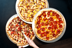 Auf dem Bild erkennt man drei sich überlappende Pizzen auf einem dunkelblauen Hintergrund. Die drei Pizzen bilden gemeinsam eine Dreieckige Form. Rechts ist eine runde Salami-Pizza zu sehen, die darüber ist eine Pizza, die mit Hühnchen und Ananas belegt zu sein scheint, und die letzte Pizza unten links ist mit Salami und noch weitere undefinierbaren Zutaten belegt. Aus dieser Pizza wird gerade von zwei Händen ein Stück hinausgeschnitten.