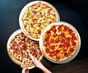 Auf dem Bild erkennt man drei sich überlappende Pizzen auf einem dunkelblauen Hintergrund. Die drei Pizzen bilden gemeinsam eine Dreieckige Form. Rechts ist eine runde Salami-Pizza zu sehen, die darüber ist eine Pizza, die mit Hühnchen und Ananas belegt zu sein scheint, und die letzte Pizza unten links ist mit Salami und noch weitere undefinierbaren Zutaten belegt. Aus dieser Pizza wird gerade von zwei Händen ein Stück hinausgeschnitten.