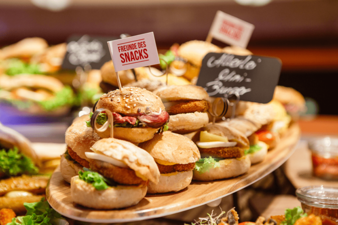Mehrere kleine Burger aus Brötchen mit Belag und Patties sind auf einem langen ovalen Holzbrett zu sehen. In den Snacks stehen kleine Fähnchen mit der Aufschrift "Freunde des Snacks".