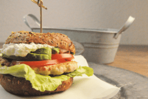 Auf dem Foto ist ein Burger zu erkennen. Die Burger Buns sind dabei aus dem proteinreichem Teig von Point of Food. Zwischen den Buns befindet sich Salt, ein Stück Hähnchen, Tomate und Gurke. Der Burger ist auf einer Serviette.