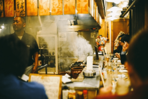 Food Impact_Delasia_Auf dem Bild ist ein kleiner asiatischer Imbisstand zu sehen, um dem wie an einer Bar leute drum sitzen und essen. Hinter dem tresen Bereich steht ein asiatischer Mann welcher am Kochen ist.
