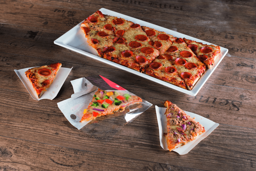 Auf dem Foto ist die GN Pizza von Point of Food abgebildet, sie in zwölf gleich große Dreiecke geteilt wird. Die GN Pizza ist auf einem weißen Teller und ist mit Salami belegt. Vor ihr sind drei Ecken auf Pappslices. Eine davon ist mit Salami belegt, die in der Mitts ist die Pizza Verdura, welche mit verschiedenen Arten Gemüse belegt ist, und der ganz rechte Point Snack ist die Thunfisch-Pizza von Point of Food. Alle Pizzen befinden sich auf einem Holztisch.