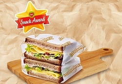 Auf dem Bild ist ein mit Schinken und Käse belegtes Sandwich zu erkennen. Beide Teile des Sandwiches liegen übereinander und sind jeweils in eine weiße Papierverpackung verkleidet. In dem Sandwich-Turm steckt ein Fähnchen von der Bel Groupe. Auf dem goldenen Stern mit rotem Band steht "Snack-Award" geschrieben. Die Bel Groupe hat nämlich dieses Jahr wieder den Snack-Award vergeben.
