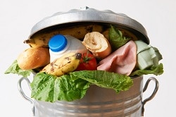 Auf dem Foto erkennt man einen silbernen Mülleimer mit Deckel, der vor Müll überquillt. Salat, Schinken, Brot, eine Banane, eine Tomate und eine Packung Milch ist als Inhalt zu erkennen. Dieses Bild weist die enorme Lebensmittelverschwendung auf.