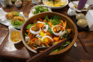Delasia_Asiatische Gemüse Bowl in einer dunkel braunen Holzschüssel und Essstäbchen serviert auf einem Tisch mit Gemüse und verschiedenen Gewürzen in kleinen weißen Schälchen. Das Gericht enthält Brokkolie, Möhren, Tempeh, Paprika und Zwiebel.
