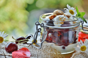 Auf dem Bild ist ein Weck-Glas zu sehen, in dem dem ein Dessert Geschichtet ist. Die untere SSchicht besteht aus einer Joghurt Creme, die zweite aus angedickten Kirschen und die odere wieder aus etwas Joghurt Creme, dekoriert mit Gänseblumen. Das Dessert ist aus einem Tisch drapiert, mit Kaffeesack, Kirschen und Gänseblumen zur Deko.