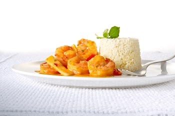 Das rote Scampi Curry von Delasia, vom Hersteller Food Impact, ist mit einer Portion Reis auf einem weißen Teller in einer hellen weißen Umgebung platziert. Auf dem Reis liegt Minze oder Basilikum zur Dekoration. Man erkennt klar und deutlich die große Garnelen in der roten Currysoße.