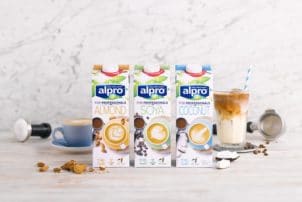 Auf dem Bild sind 3 Milchalternativen von Alpro for Professionals in Tetra Paks zu sehen. Dabei handelt es sich um Mandel-, Soja- und Kokosnussmilch. Neben den Tetra Paks sind ein Kaffee in einer Tasse und ein Milchshake in einem Glas zu sehen.