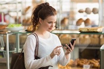 Eine Frau in einer Bäckerei, die ein Smartphone in der Hand hält. Um sie rum befinden sich Theken mit Backwaren.