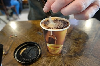 Ein Kaffee in einem to go Becher, in dem gerade Zucker gefüllt wird.