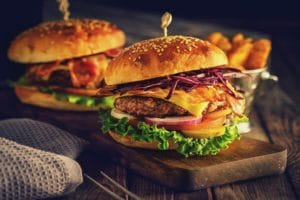Auf dem Bild sind zwei Burger zu sehen, die sich auf einer Holzplatte auf einem Holztisch befinden. Der Burger ist mit einer Bulette, Käse, Speck, Gurken, Zwiebeln, Tomaten und Salatblättern belegt. Im Hintergrund ist eine Schale mit Pommes zu erkennen.
