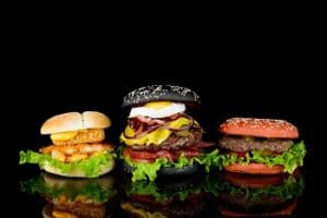 Auf dem Bild erkennt man farbige Burger vor einem schwarzen Hintergrund. Der rechte Burger hat rote Burgerbrötchen, ist mit einer Bulette, einem Salatblatt und Tomaten belegt. Der Burger in der Mitte hat schwarzes Burgerbrot und ist mit Ei, Speck, Käse, Zwiebeln, Tomaten und Salat belegt. Der linke Burger hat normales Burgerbrot und ist mit Hühnchen und Salat belegt.