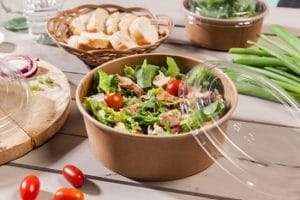 Auf dem Bild ist ein Salat in einer Kartonschale von Bionatic zu sehen. Der Salat besteht aus Rucola, Salatblättern, Tomaten und Hühnchen. Auf der Schale liegt ein aus Kunststoff bestehender Deckel. Im Hintergrund sind eine Schale mit Brot, so wie verschiedenstes Gemüse zu sehen.