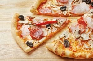 Ein Stück einer Pizza belegt mit Salami, Schinken, einer Chilischote und Pilzen auf einem Holzbrett.